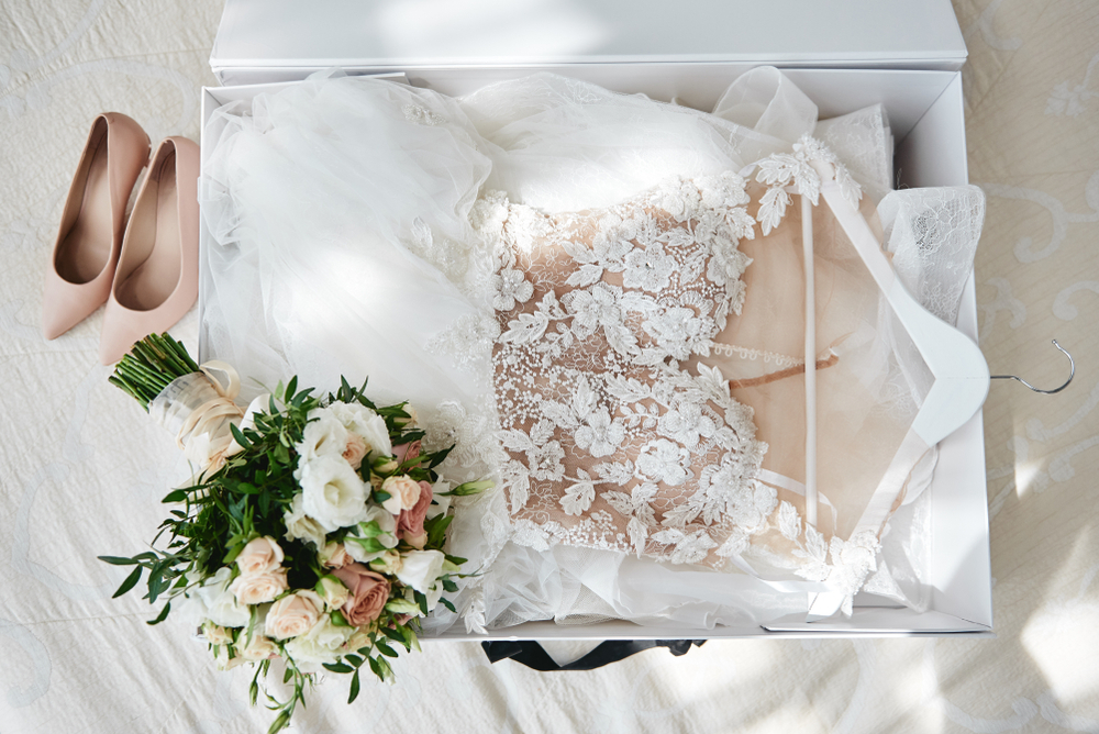 Luxury wedding dress in white box, beige women's shoes and bridal bouquet on bed, copy space. Bridal morning preparations