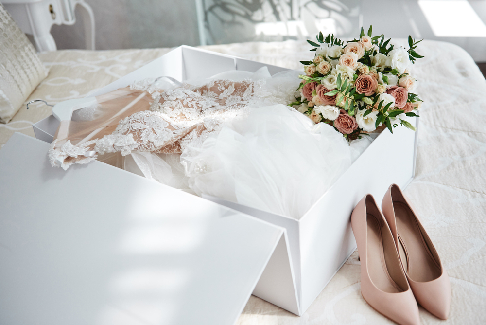 Luxury wedding dress in white box, beige women's shoes and bridal bouquet on bed, copy space. Bridal morning preparations. Wedding concept
