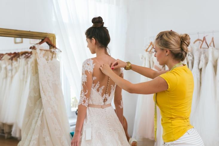 dressmaker helping the bride to put her wedding dress on in clothes shop.