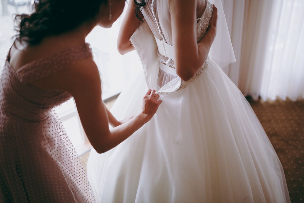 Bridesmaid helping slender bride lacing her wedding white dress, buttoning on delicate lace pattern with fluffy skirt on waist. Morning bridal preparation details newlyweds.